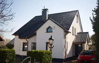 Haus mit steilem Satteldach, weißer Fassade, Gaube und rotem Auto neben dem Eingang