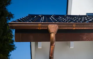 Nahaufnahme eines Kupferdachrinnensystems mit Fallrohr an einem weißen Haus unter blauem Himmel
