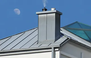 Dach mit grauem Schornstein, aus dem Rauch aufsteigt, und sichtbarem Mond am blauen Himmel